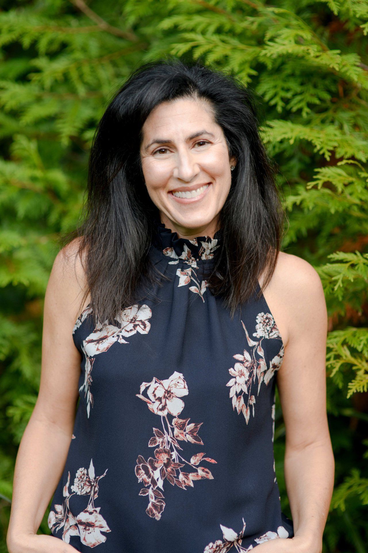 A woman in a floral dress is smiling in front of a tree.