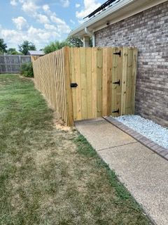Wooden privacy fence gate beside a sidewalk and lawn, with gravel along the house foundation.