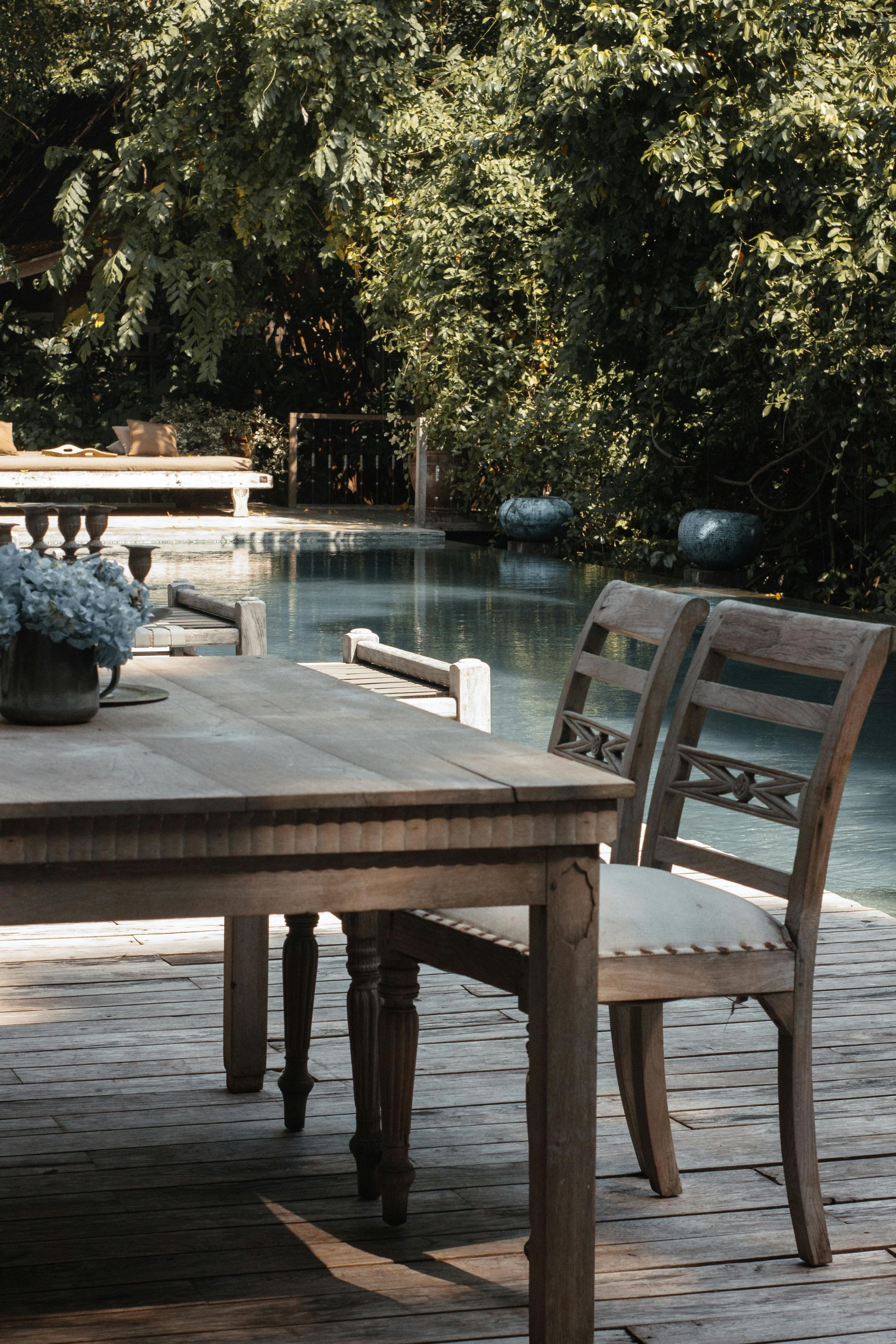 Wooden patio table and chair beside a sunlit pool surrounded by trees