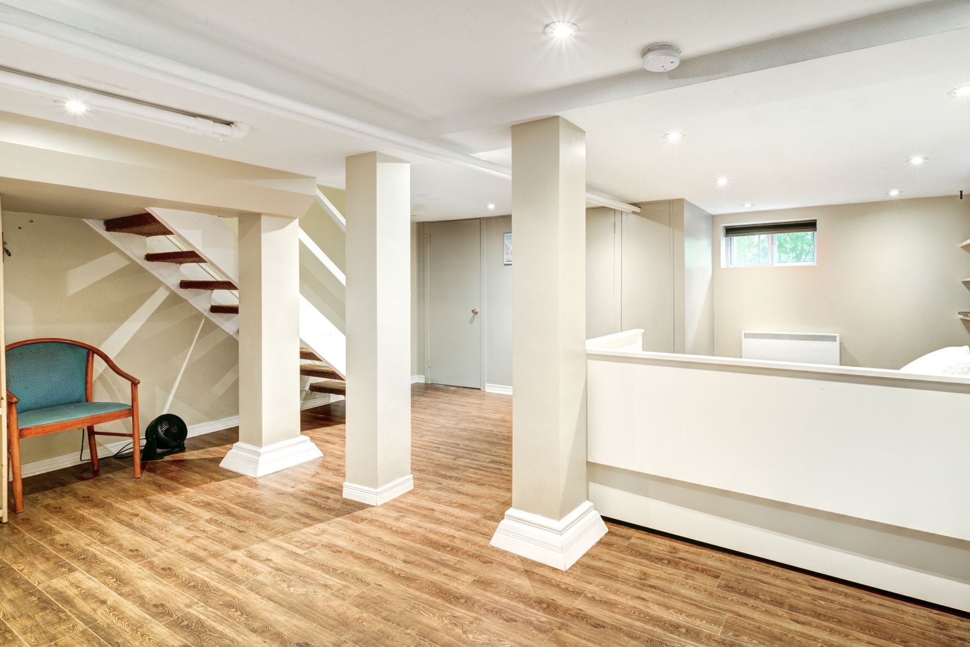Bright basement room with wood floors, white columns, stairs, and a half-wall bar area