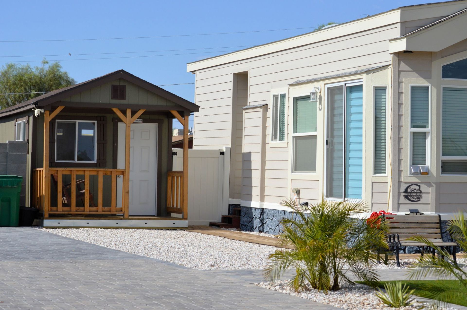 Small wooden porch beside a white mobile home on a sunny day, with gravel and a patio area.