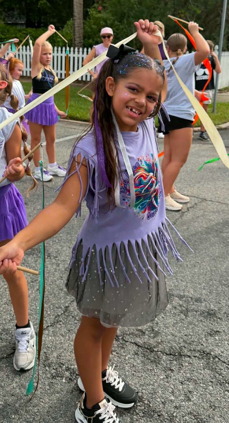 Smiling girl in purple fringe top and gray skirt holding ribbon streamers at an outdoor event