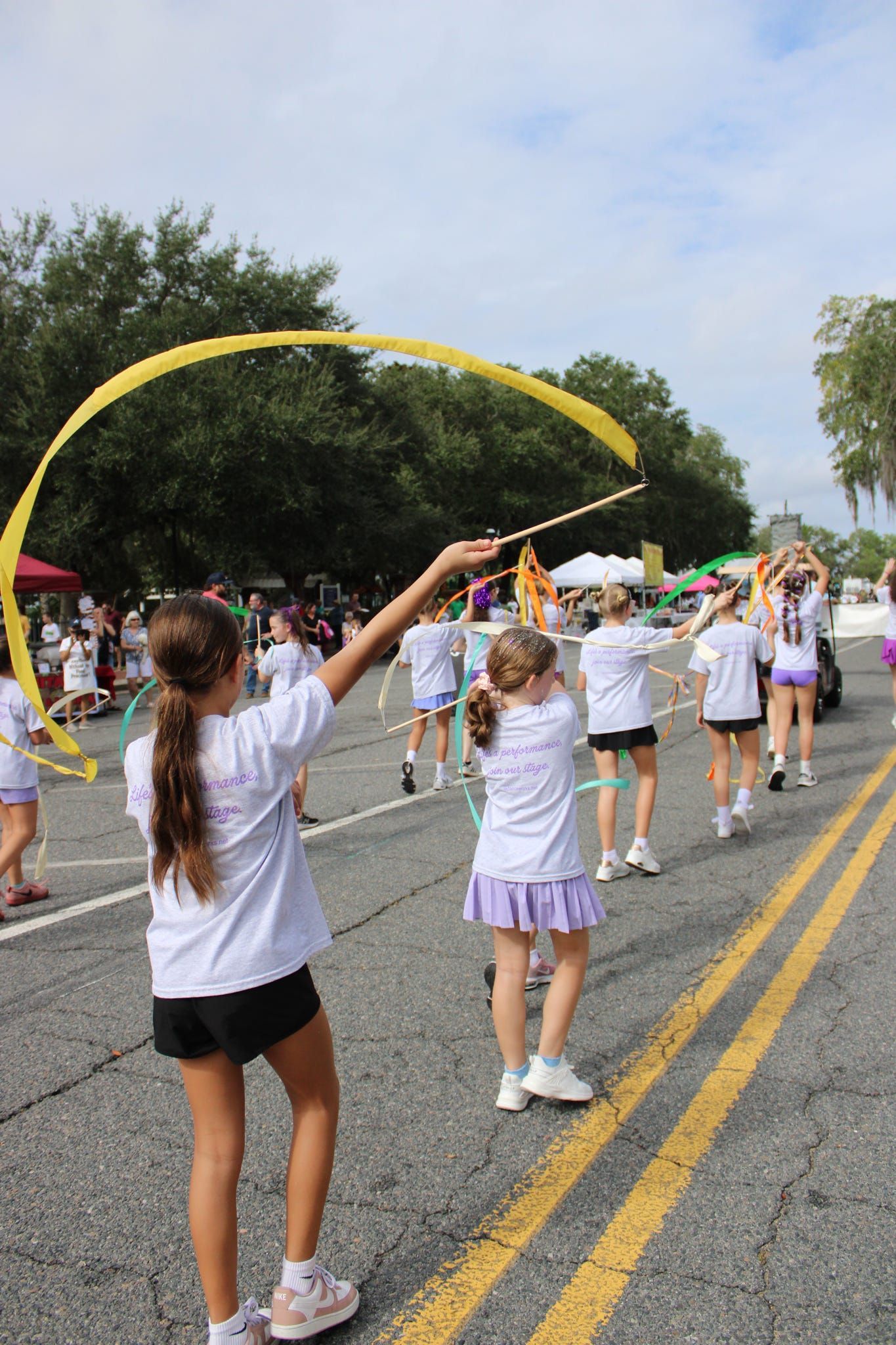 Children in white shirts practice with yellow ribbons on a street during a parade rehearsal