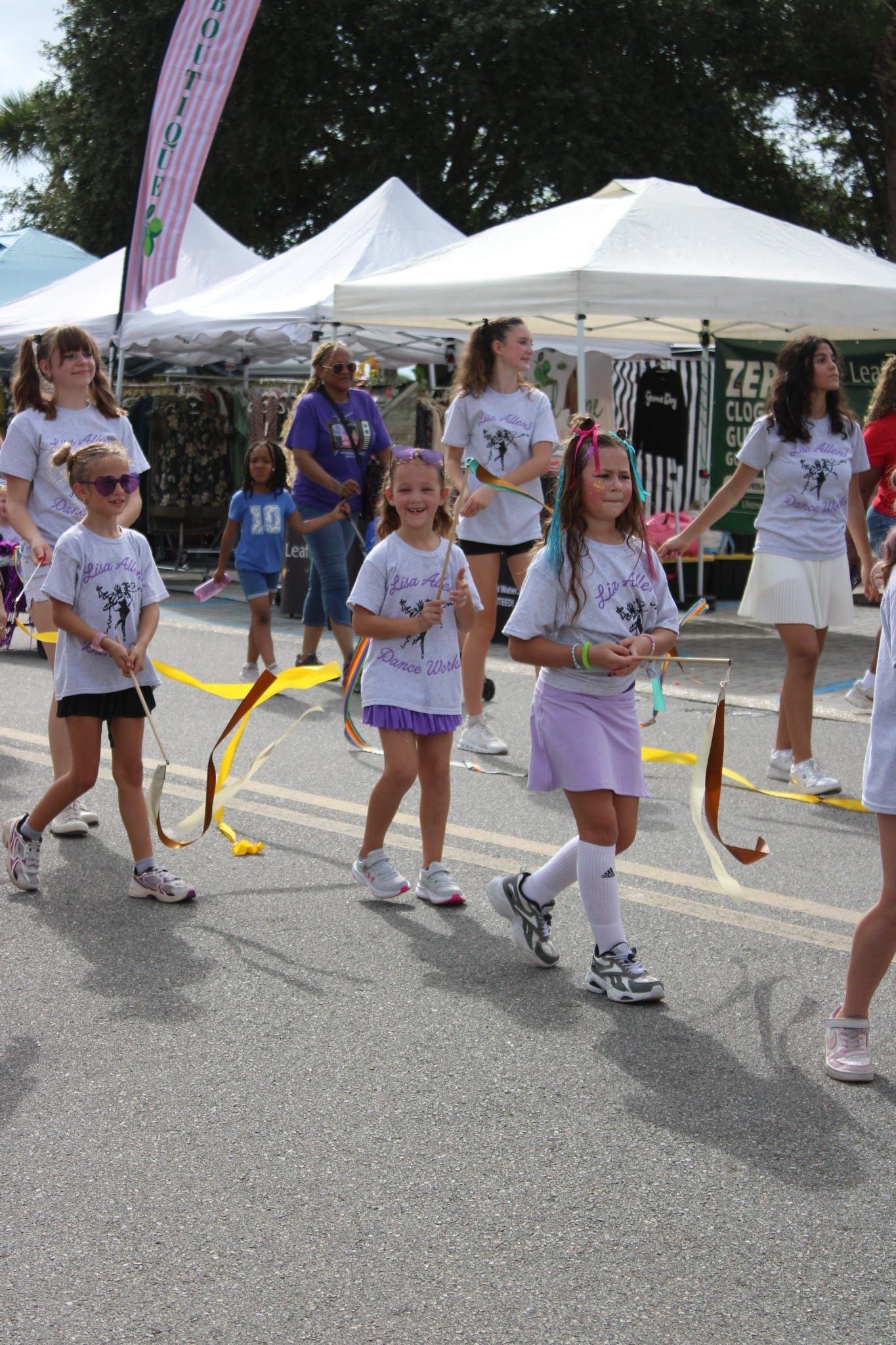 Children in white shirts and purple shorts dancing in a street parade near white tents