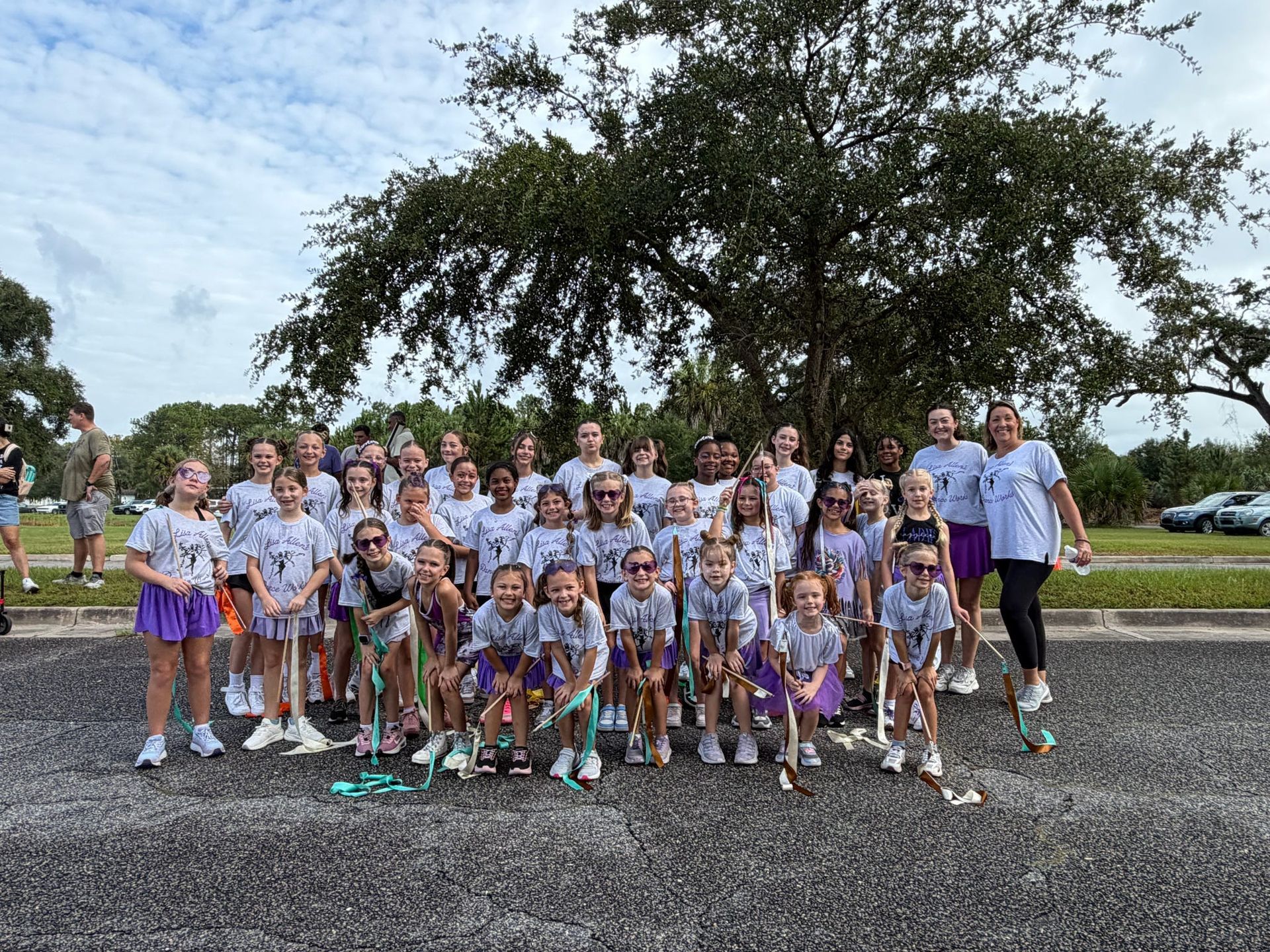 Group of children and adults posing outdoors in matching purple shirts on a paved area under a tree