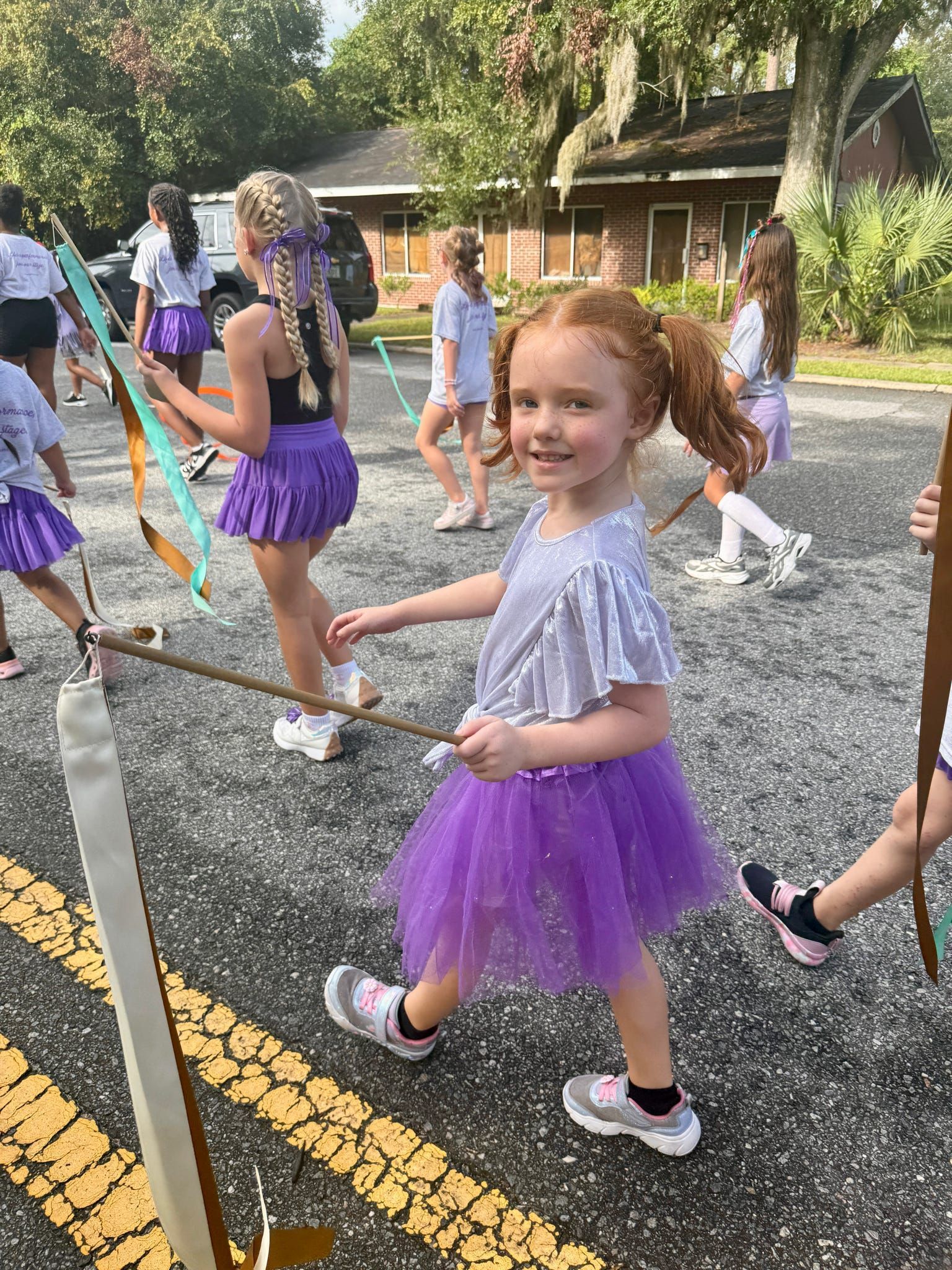 Children in purple tutus dancing outdoors on a gravel path in a park-like setting