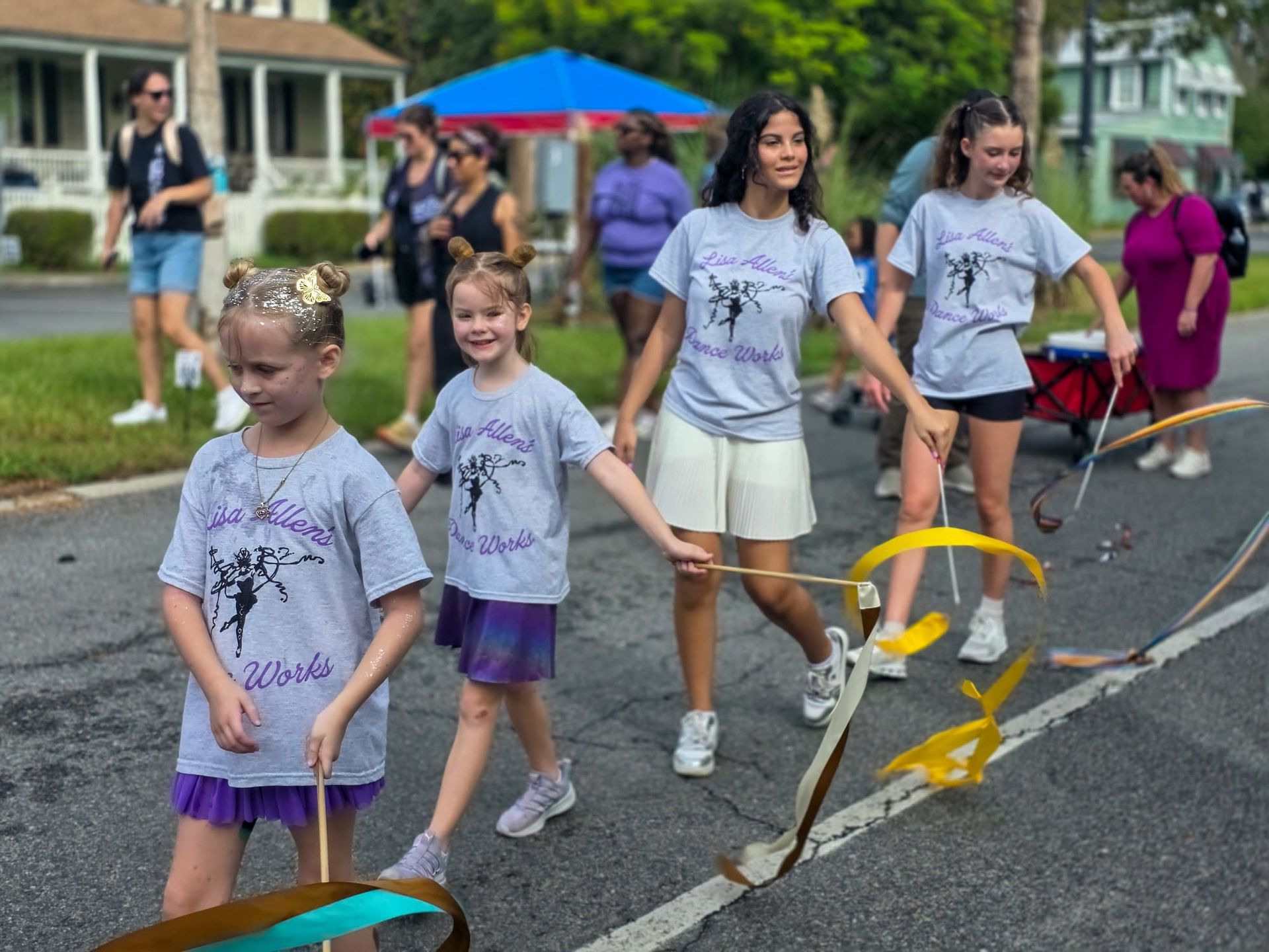Children walking together in a neighborhood parade, wearing matching T-shirts and carrying a small yellow ribbon decoration.