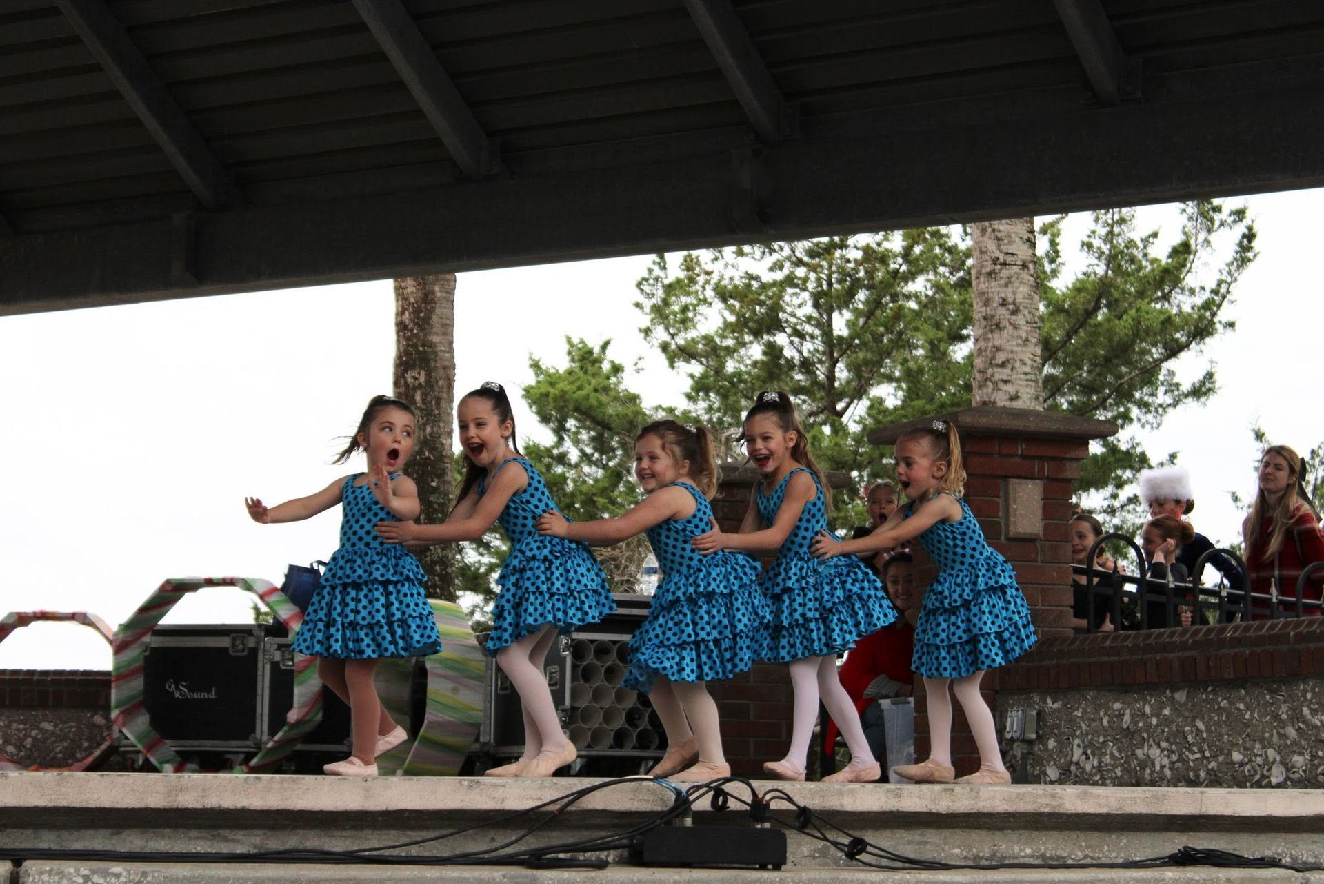Dancers in blue dresses perform on an outdoor stage with musicians in the background