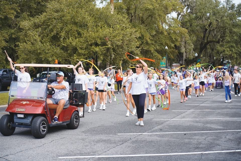 Parade participants in white shirts walk down a street, with a red golf cart leading the group.