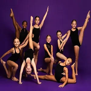 Group of young dancers posing in black leotards against a purple backdrop.