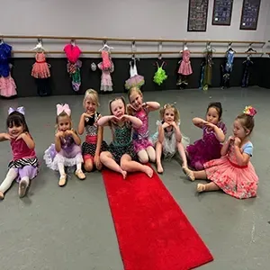 Children in colorful dance costumes sitting on a red carpet in a ballet studio