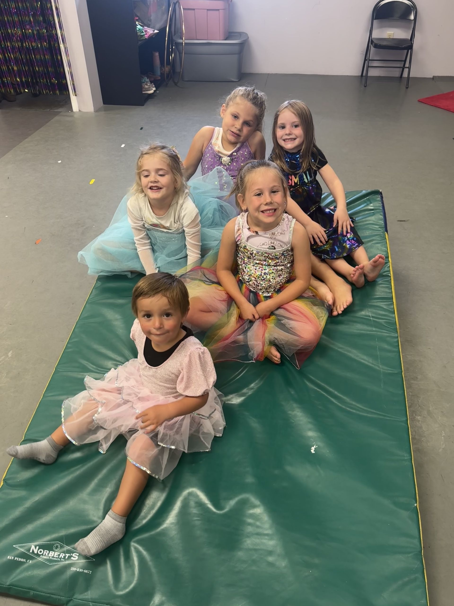 Children in ballet outfits sitting on the floor in a dance studio, with a red carpet and tutus hanging behind them
