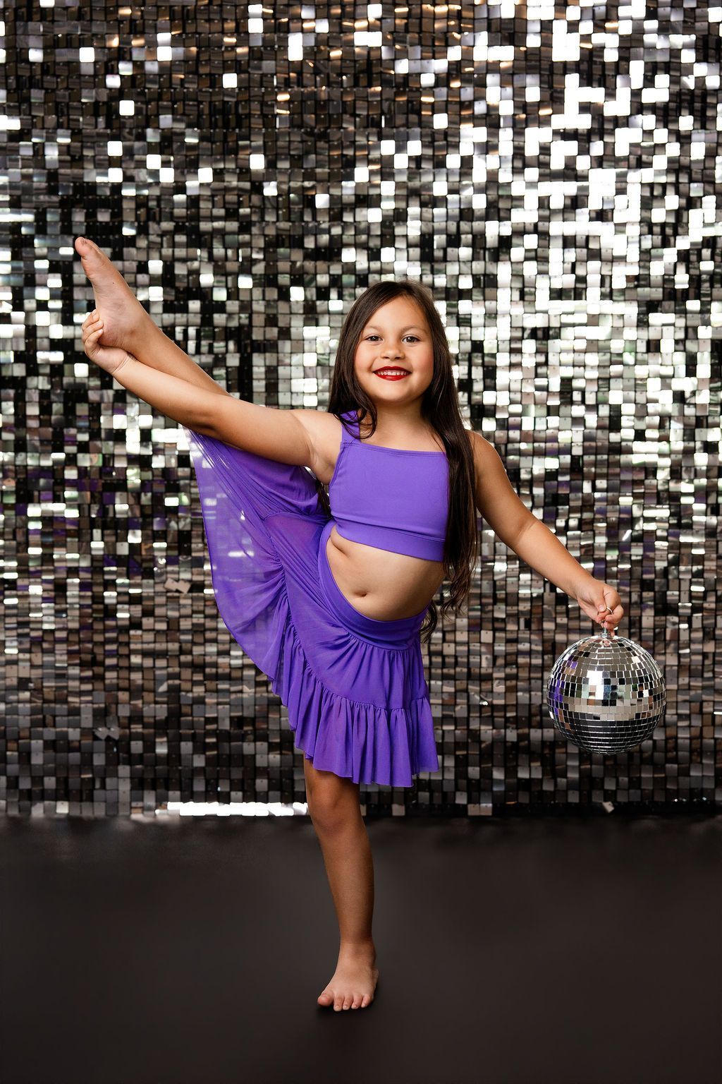 Child posing in a black leotard against a purple confetti backdrop, smiling with one arm raised.