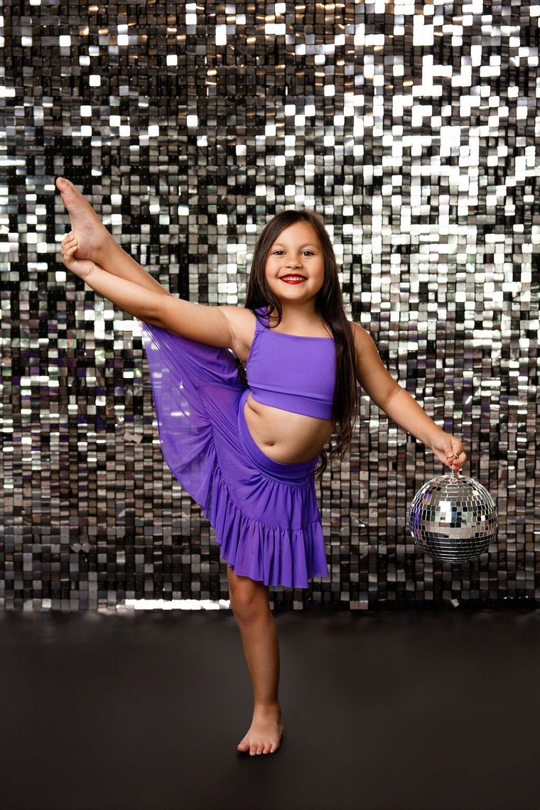 Child posing in a black leotard against a purple confetti backdrop, smiling with one arm raised.