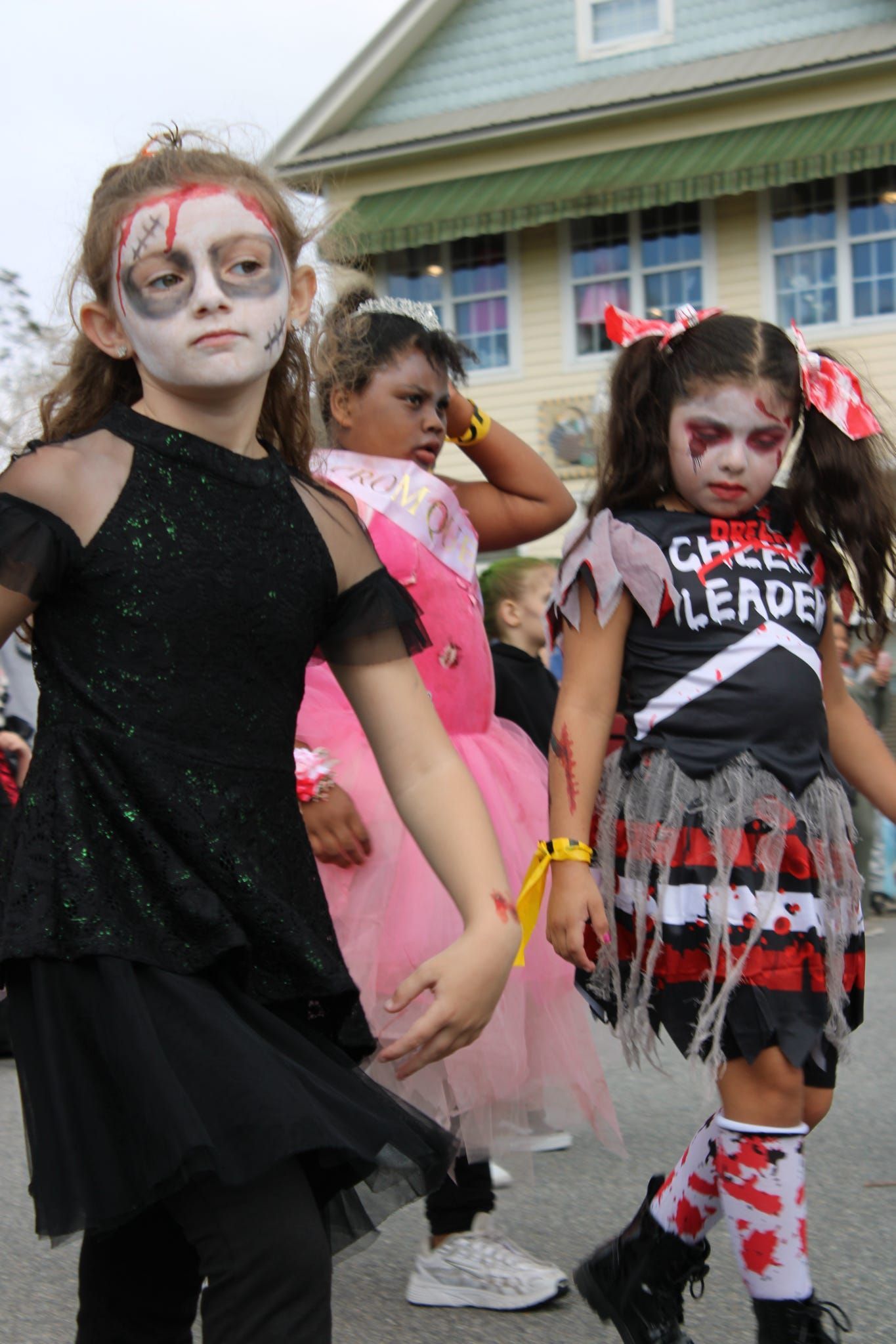 Children in Halloween costumes walking outdoors, one with face paint and one in a black “Cheerleader” outfit.