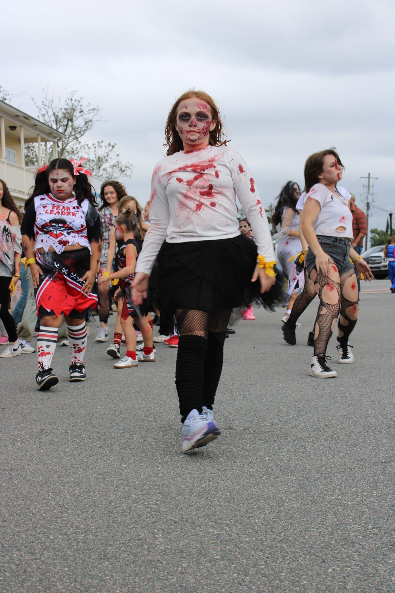 People in zombie makeup walking in a street parade, with a girl in a white shirt and black skirt in front