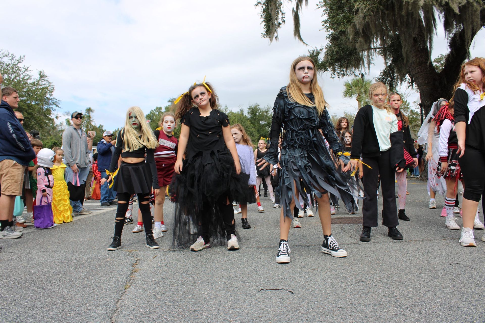 Costumed crowd walking on a street, with several people in dark outfits in the foreground and palm trees behind them