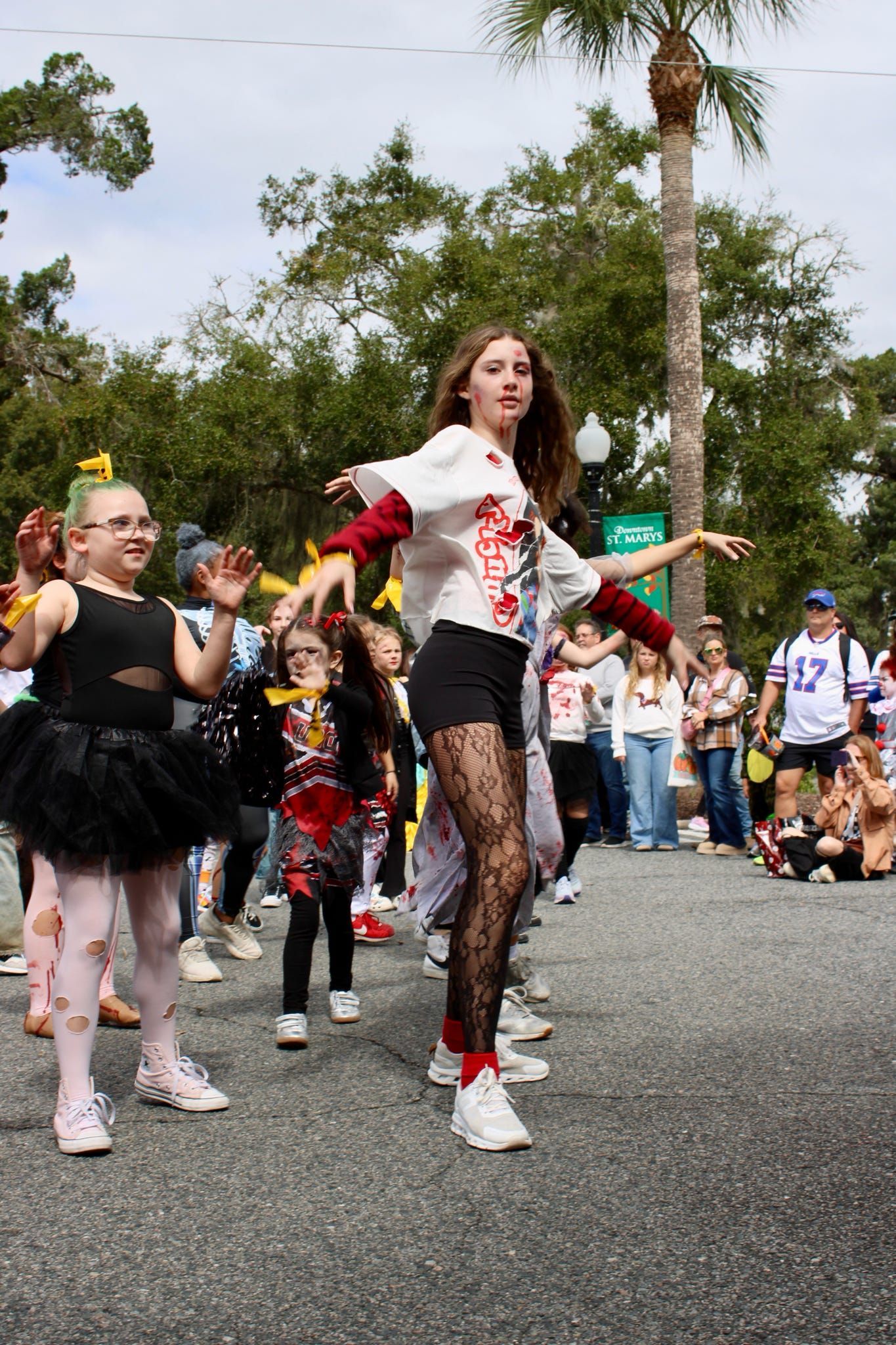 Costumed dancers perform in a street parade, with spectators and palm trees in the background