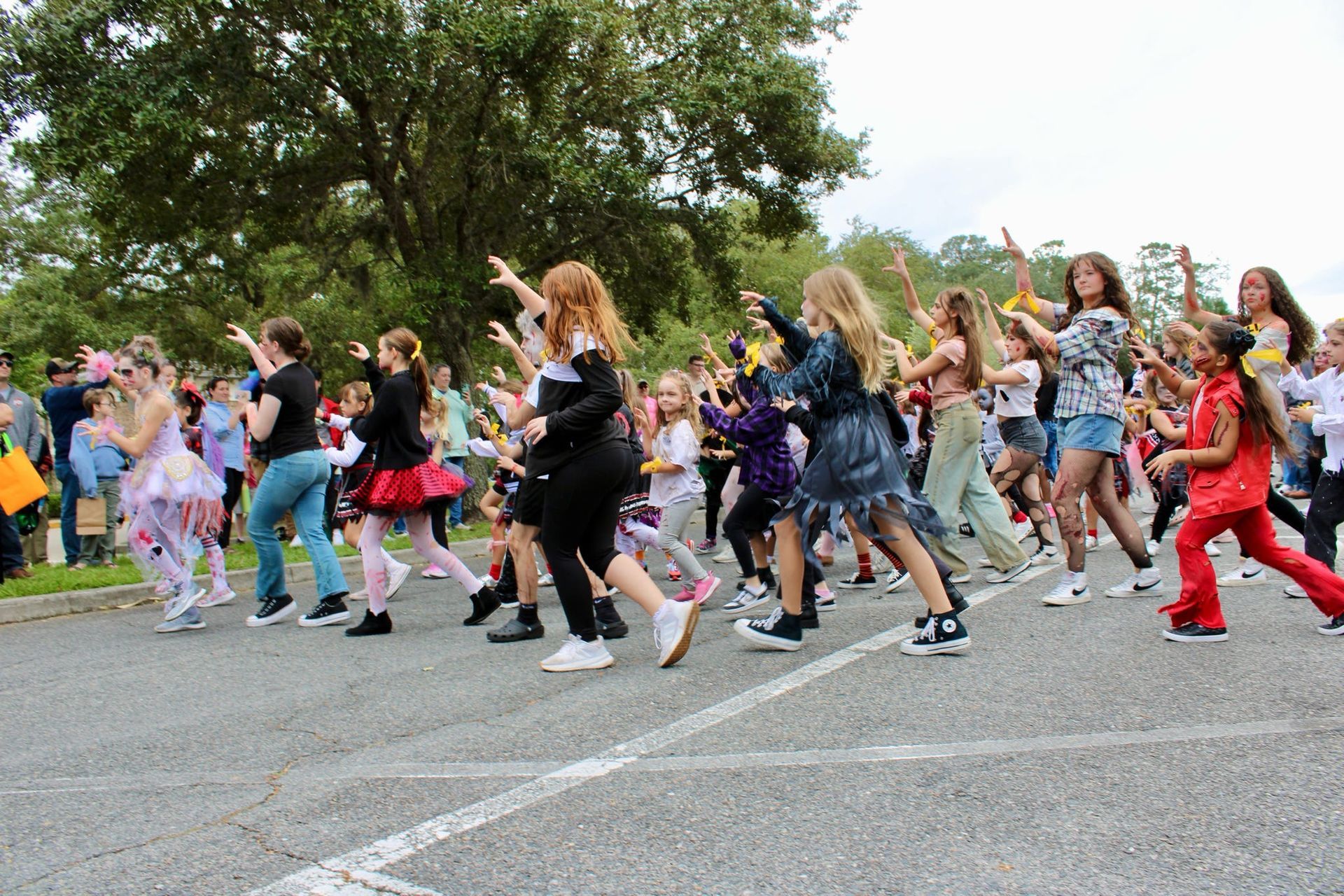 Crowd of people dancing in a parking lot, wearing colorful outfits with trees in the background