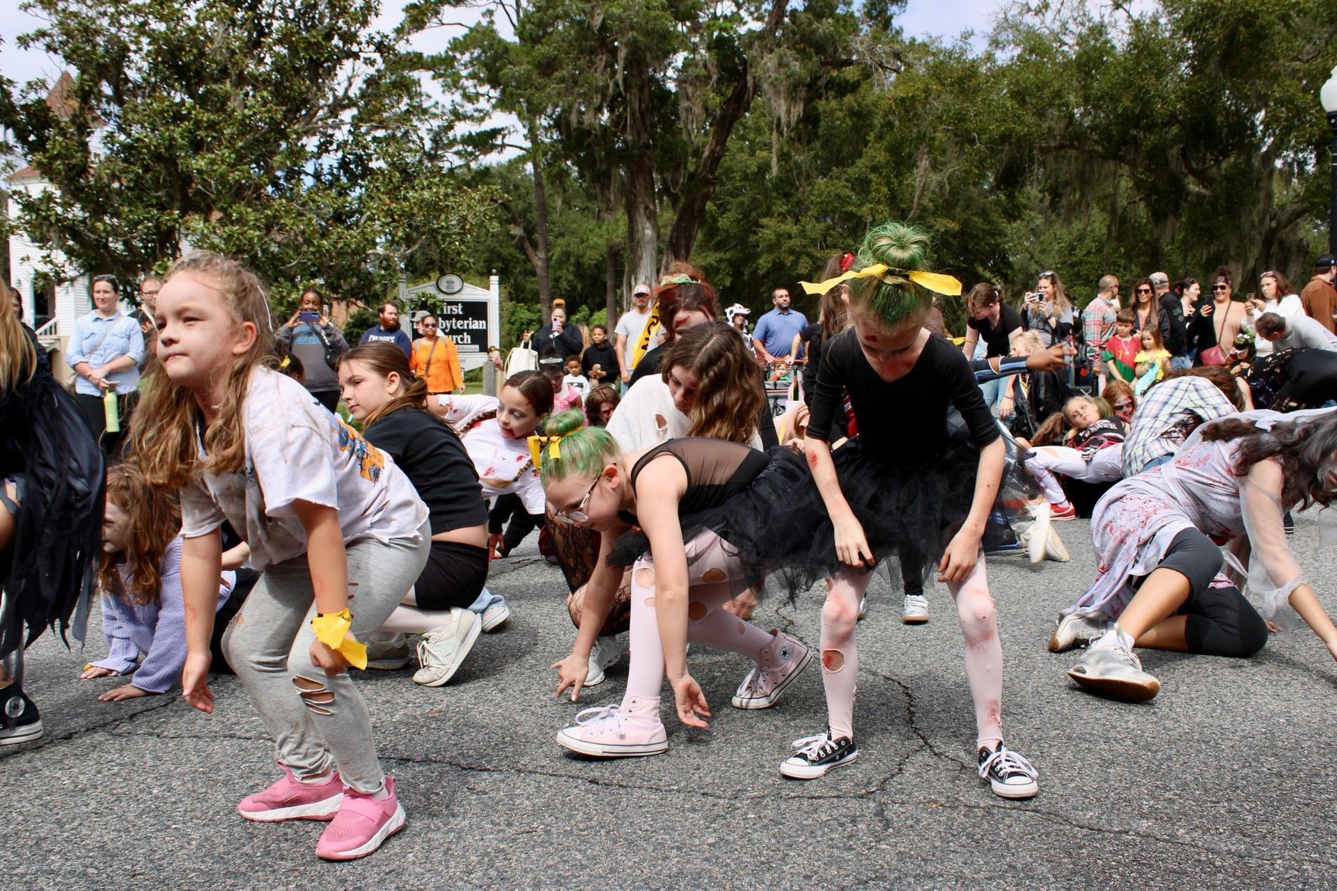 Children stretching outdoors in a group exercise, wearing colorful sneakers on a paved area with trees and spectators.