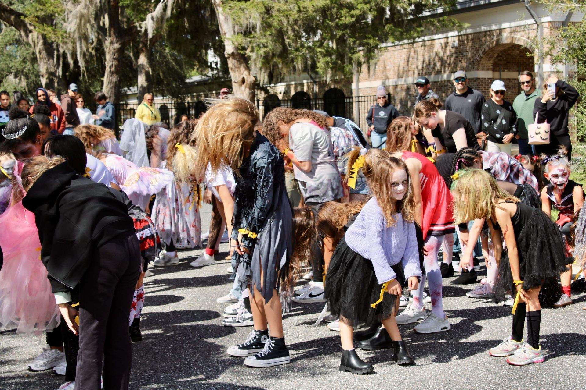 Crowd of people in costumes dancing outdoors at a street festival with trees and a building behind them