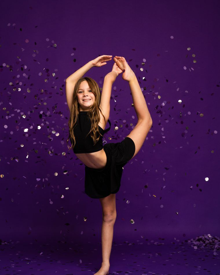 Young dancer in black dress striking a high leg pose against a purple backdrop with confetti