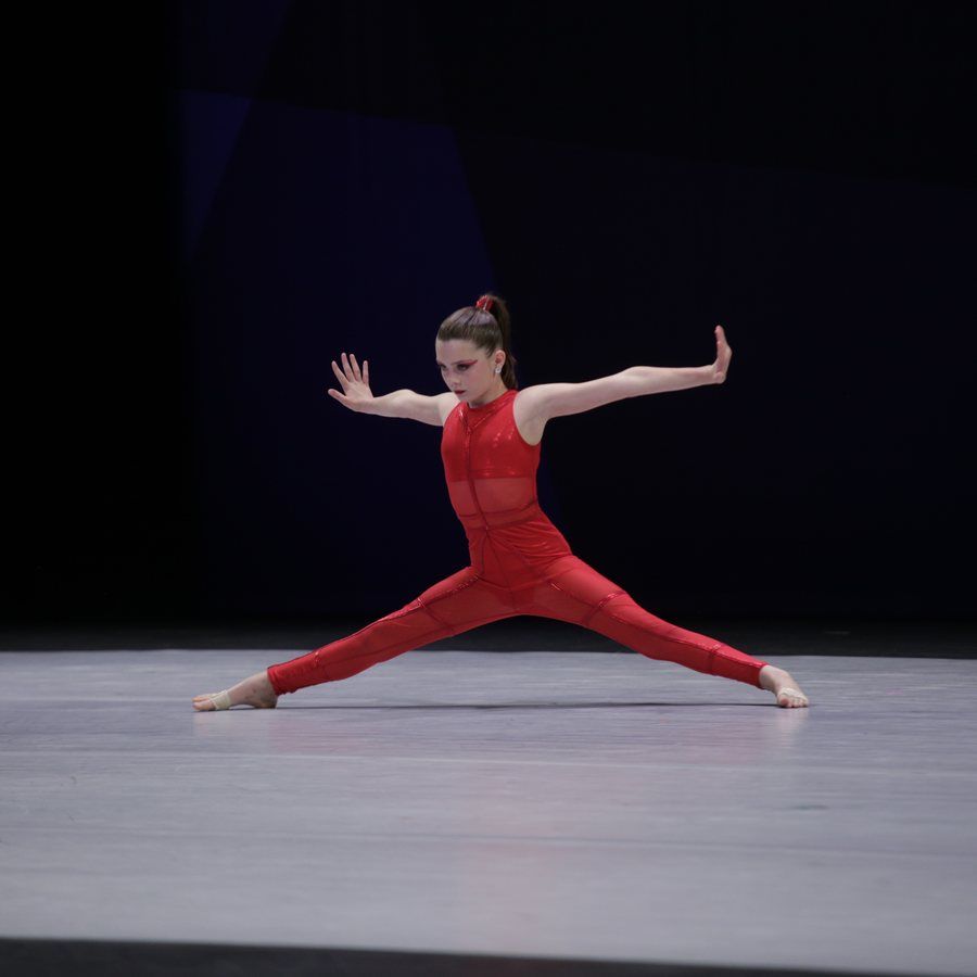 Dancer in a red outfit holds a wide split pose on a stage under dark lighting.