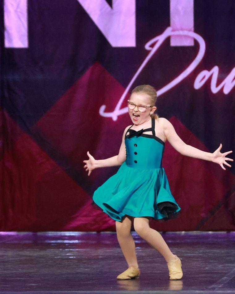 Young dancer in teal dress performing on stage with arms outstretched, smiling under stage lights.