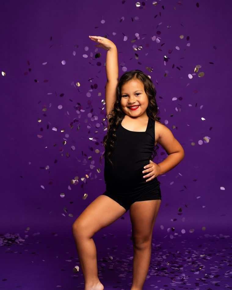 Child posing in a black leotard against a purple confetti backdrop, smiling with one arm raised.