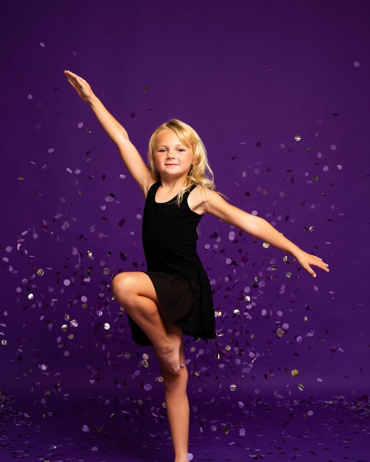 Young dancer in black outfit posing with one leg raised against a purple background