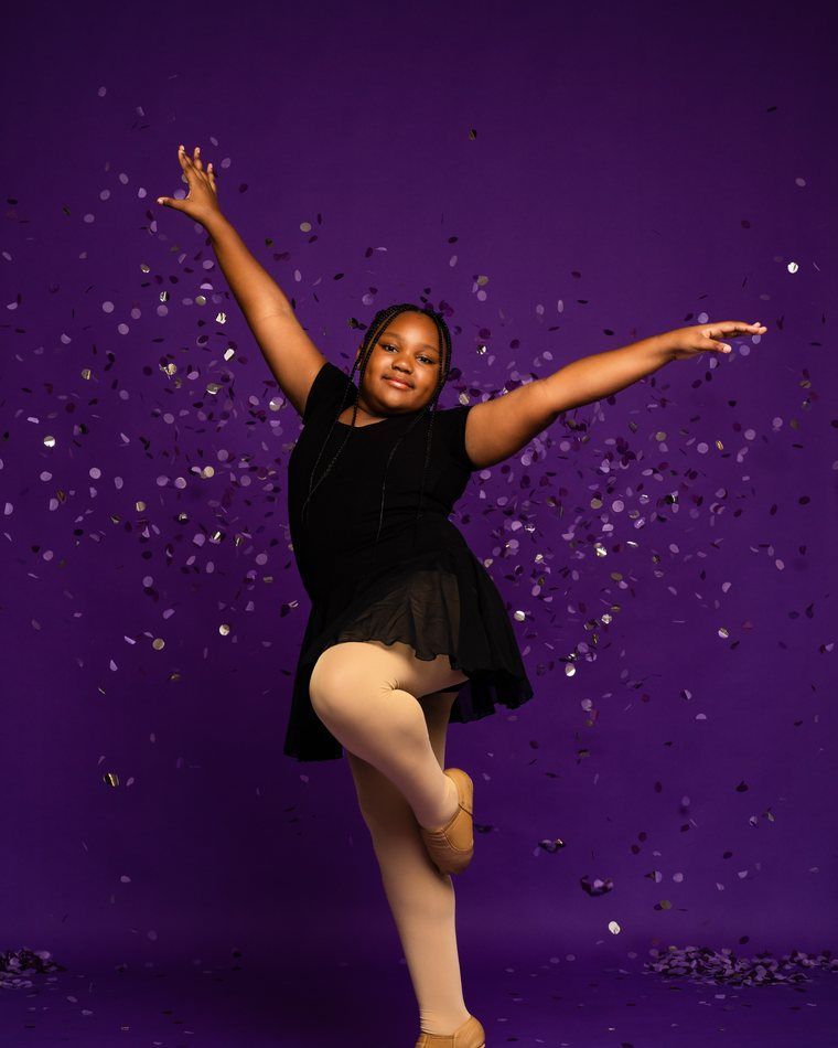 Dancer in a black dress and tights poses with arms raised against a purple confetti backdrop.
