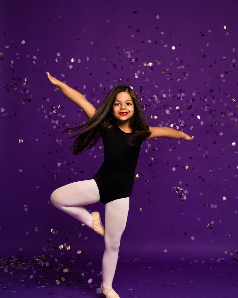 Girl in black leotard and white tights posing ballet-style against a purple confetti backdrop.