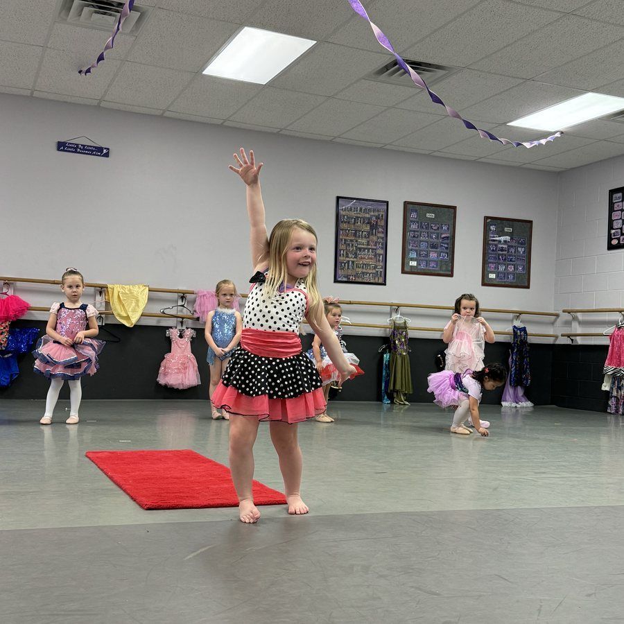 Young child dancing with one arm raised in a decorated studio, with other children in the background.