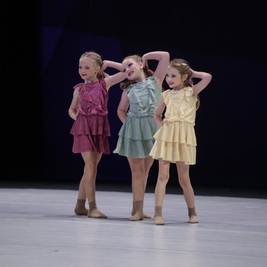 Three young dancers in colorful dresses pose on a stage with hands behind their heads.
