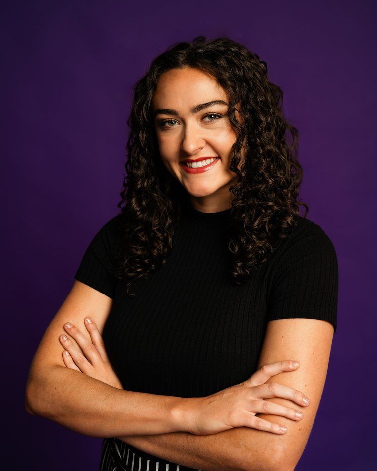 Smiling woman with curly hair and crossed arms against a purple background