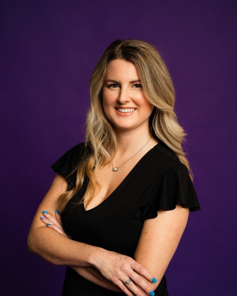Smiling person in a black dress, standing against a purple background with arms crossed.