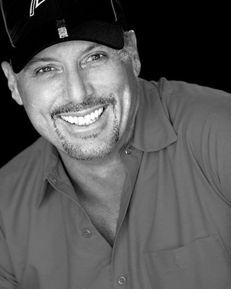 Smiling man in a black cap and red shirt, shown in black and white against a dark background