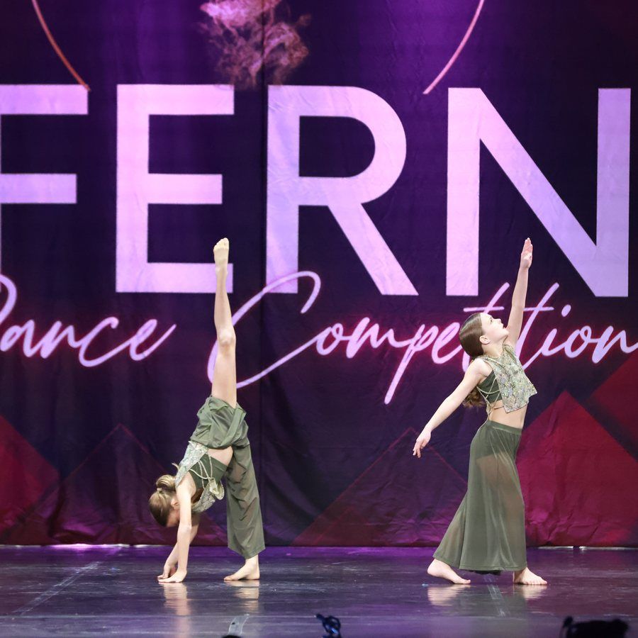 Two dancers in green costumes perform onstage before a large purple dance competition backdrop.