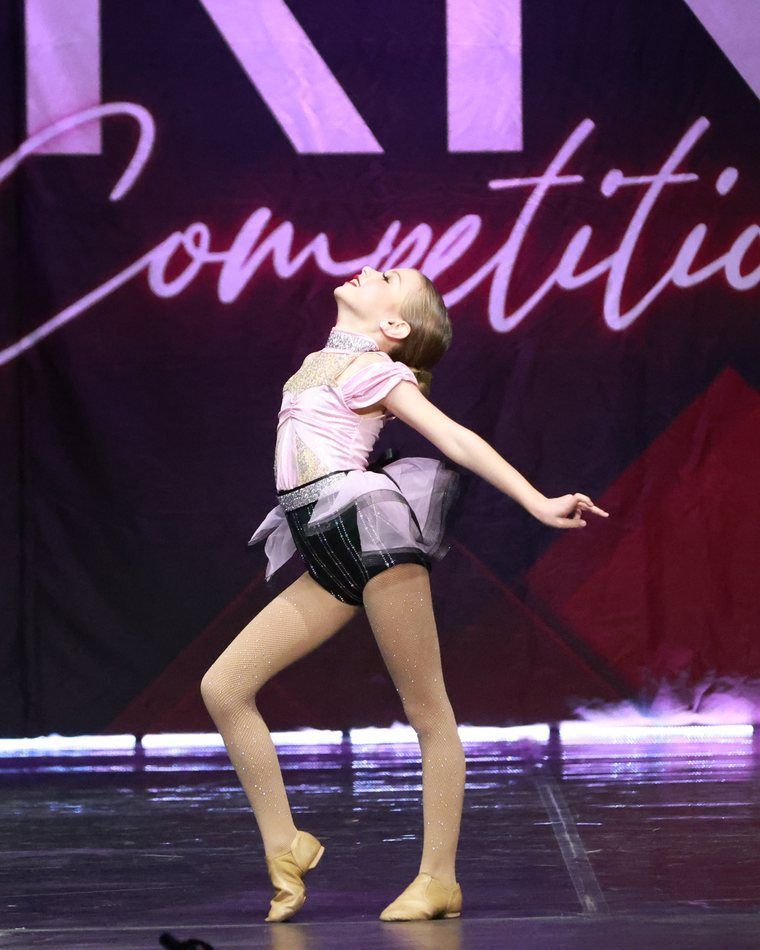 Ballet dancer in pink costume poses onstage under purple lighting with a competition banner behind her