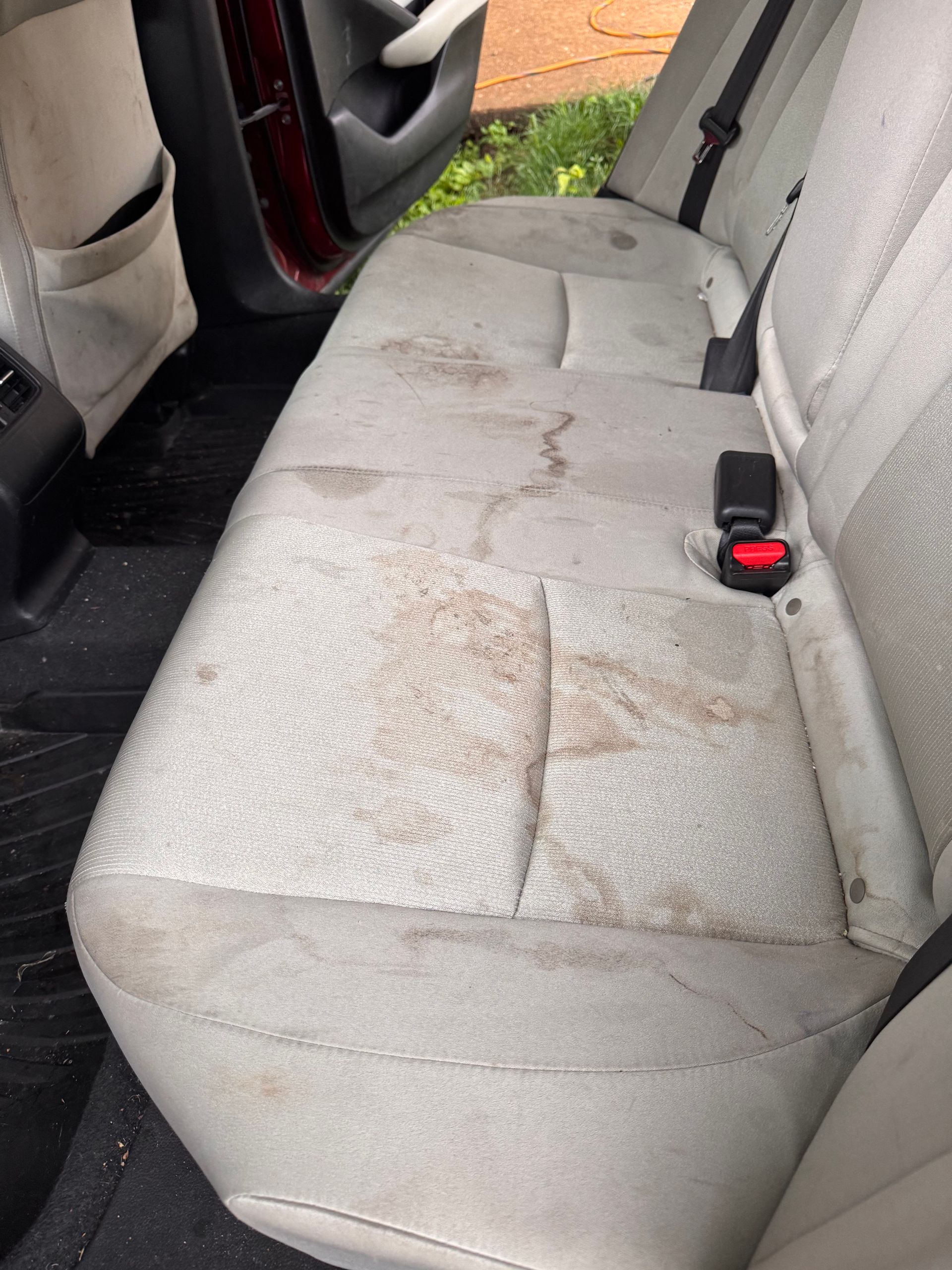 Dirty beige back seat of a car with stains and a red seatbelt buckle visible