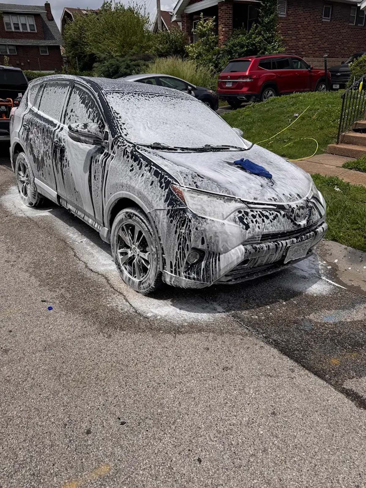 White car covered in foam at a driveway car wash, with soap suds on the road around it