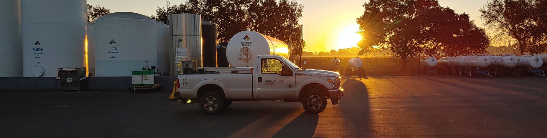 A white pickup truck with a large tank drives near storage tanks at sunrise.