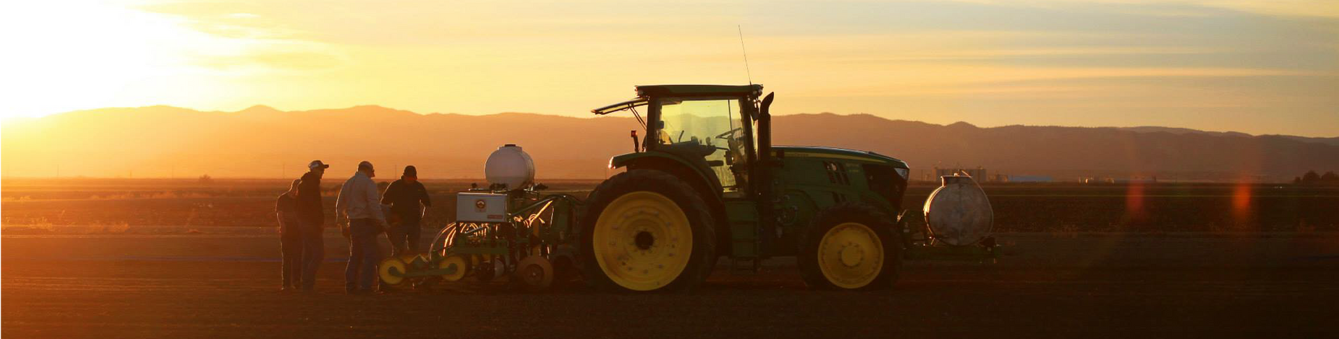 Tractor and farm equipment in a field at sunset, with silhouetted figures nearby.