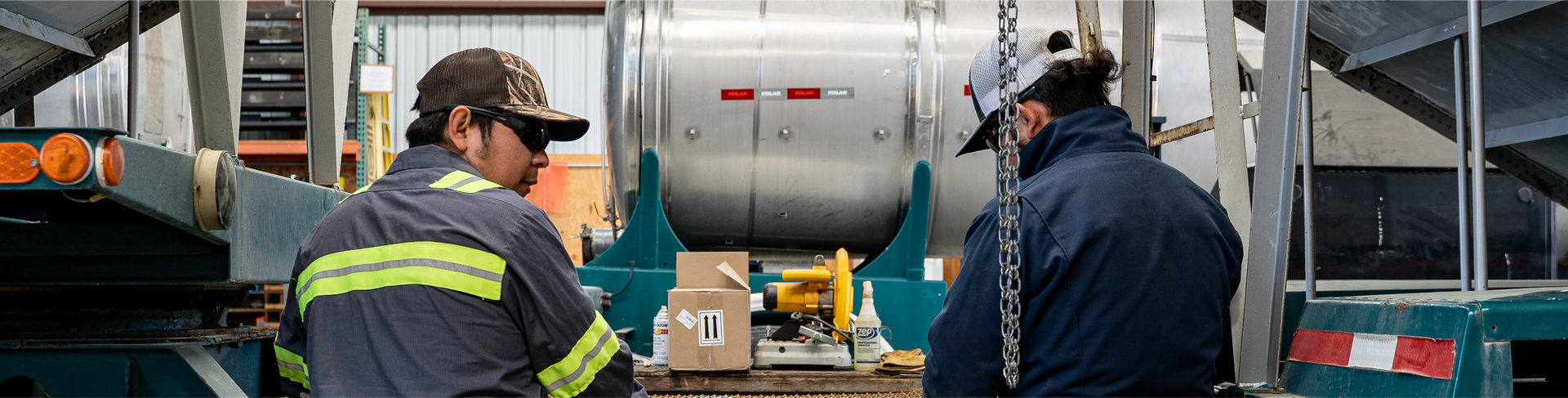 Two workers near a metal cylinder. One wears a cap and hi-vis stripes. Boxes sit between them.