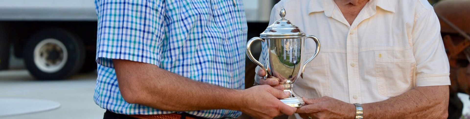Two men holding a silver trophy outdoors. One wears a blue checked shirt, the other a cream shirt.