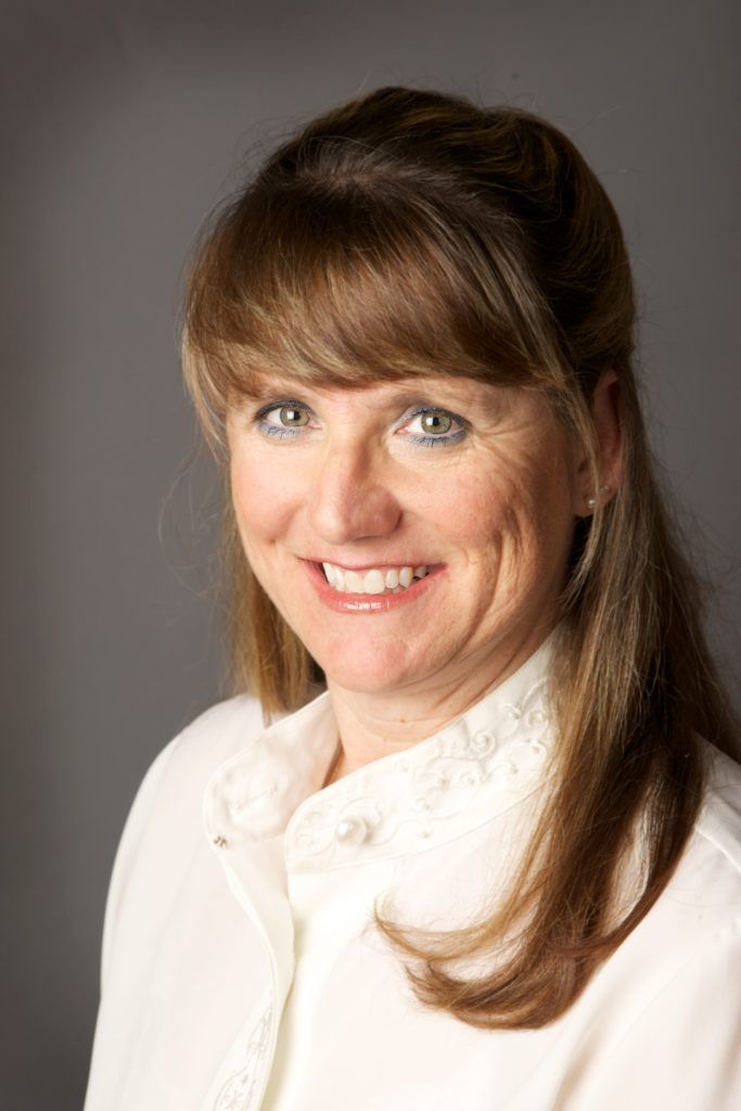 Woman with light brown hair, smiling, wearing a white shirt against a gray backdrop.