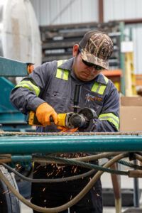 Worker in gray jumpsuit using a grinder, sparks flying. Outdoors, working on metal with safety gear.