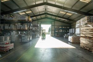 Warehouse interior with a forklift, shelves of boxes, and pallets of goods. Sunlight streams through the open doorway.