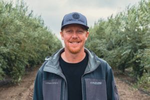 Man wearing a baseball cap and jacket smiles outdoors in an orchard.