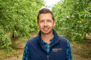 Man in a vest and plaid shirt smiles in front of an orchard.